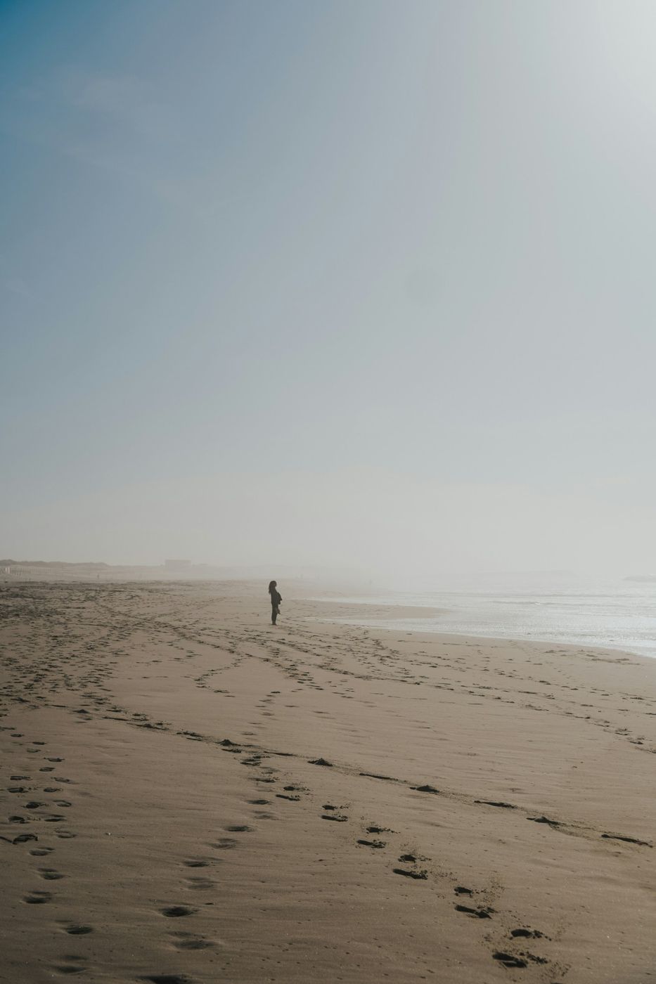 A lone figure standing on a wide, misty beach, footprints in the sand
