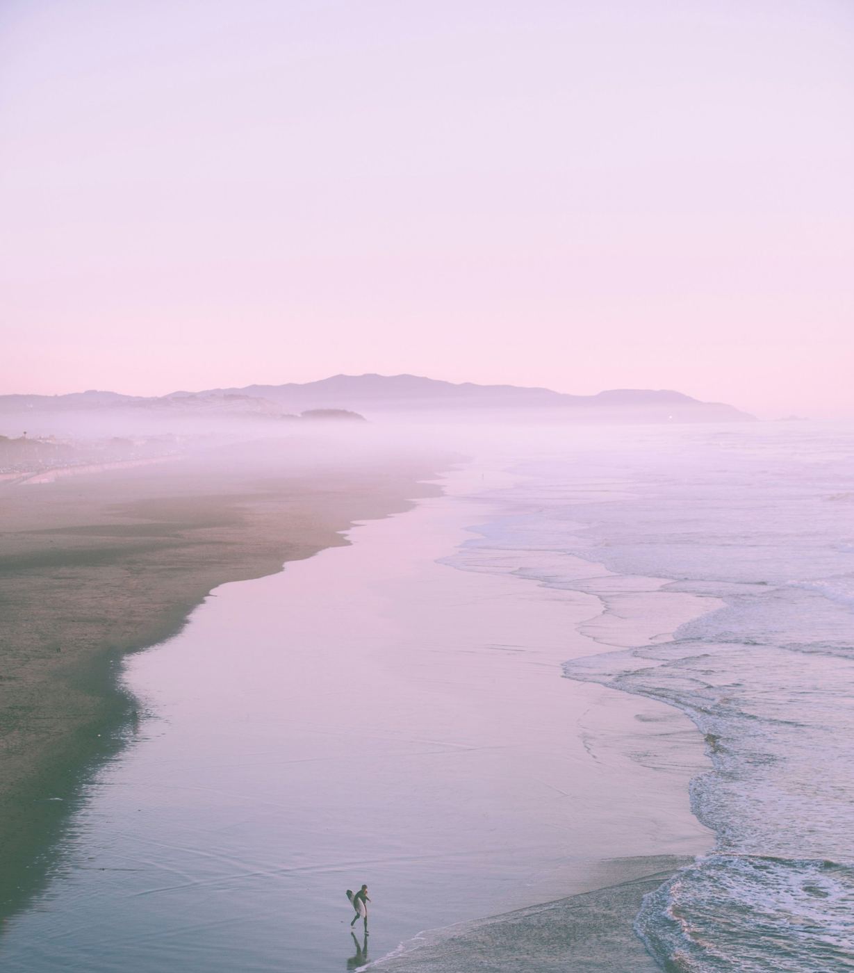 A curving shoreline in pink mist, a lone surfer small at the water's edge